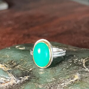 May include: A silver ring with a large, oval, bright green gemstone. The gemstone is set in a gold bezel. The ring band has a textured design. The ring is displayed on a green, textured surface.