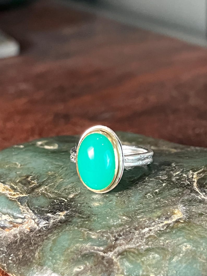 May include: A silver ring with a large, oval, bright green gemstone. The gemstone is set in a gold bezel. The ring band has a textured design. The ring is displayed on a green, textured surface.