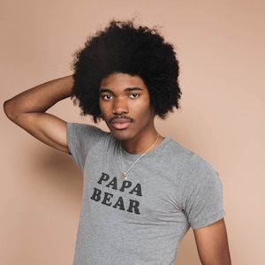 以下が含まれることがあります： グレーのTシャツを着た男性。Tシャツには「パパベア」の文字がプリントされています。彼は茶色のズボンを履いており、小さなペンダントのついたシルバーネックレスをしています。