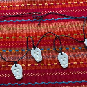 May include: A string of five white ceramic skull charms hanging from black string on a red and blue striped fabric background.