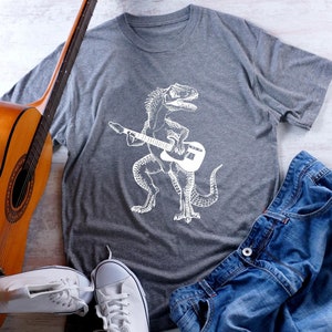 May include: A gray t-shirt with a white graphic of a dinosaur playing a guitar. The shirt is laid out on a white wooden surface with a guitar, jeans, and white sneakers.