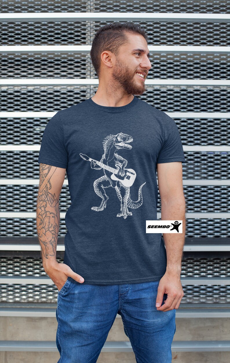 May include: A navy blue t-shirt with a white graphic of a dinosaur playing a guitar. The t-shirt has the text "SEEMBO" on the bottom right corner.