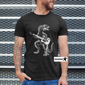 May include: A dark heather grey t-shirt featuring a white graphic of a dinosaur playing a guitar. The t-shirt has the text "Seembo" on the bottom right corner.