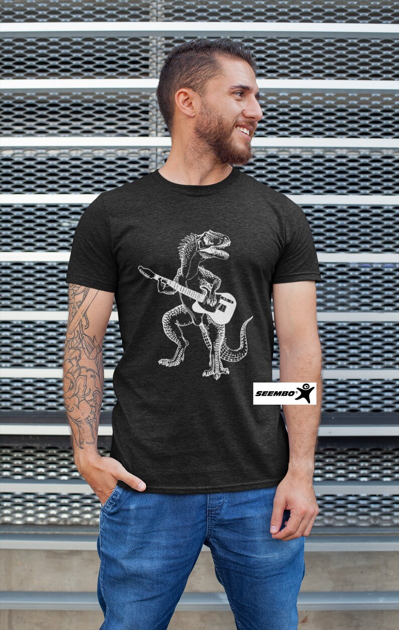 May include: A dark heather grey t-shirt featuring a white graphic of a dinosaur playing a guitar. The t-shirt has the text "Seembo" on the bottom right corner.