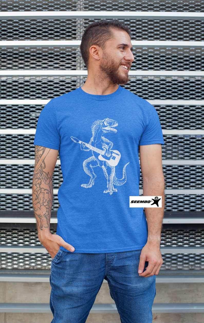 May include: A royal blue t-shirt with a white graphic of a dinosaur playing a guitar. The dinosaur is standing on its hind legs and holding a guitar in its front paws. The t-shirt has the word "SEEMBO" printed on the bottom right corner.