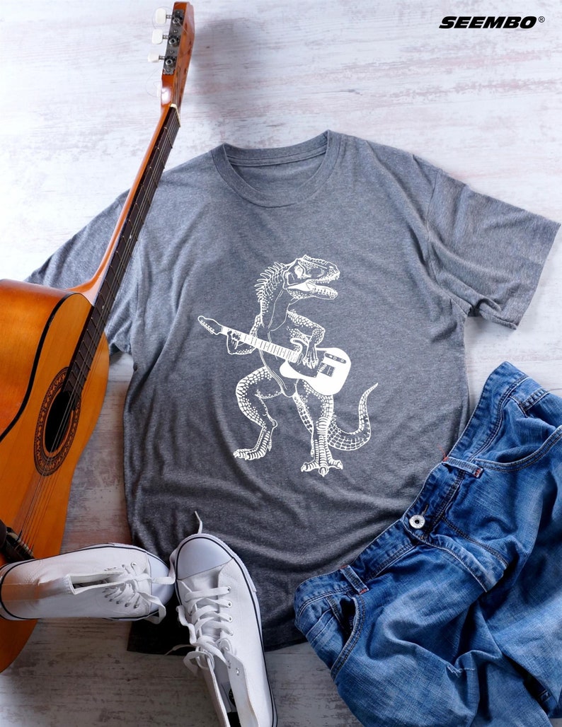 May include: A gray t-shirt with a white graphic of a dinosaur playing a guitar. The shirt is laid out on a white wooden surface with a guitar, jeans, and white sneakers.