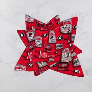 May include: Set of four red and grey pot holders featuring a University of Georgia Bulldogs theme. The pot holders have a repeated pattern of the team's logo, mascot, and the words "Georgia Bulldogs".