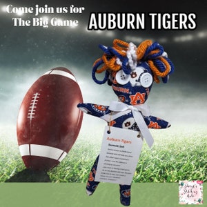May include: An Auburn Tigers Dammit Doll with orange, blue, and white yarn hair and button eyes. The doll is holding a brown football. The text "Come join us for The Big Game" and "AUBURN TIGERS" are visible.