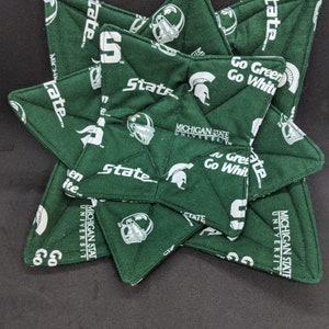 Puede incluir: Una pila de salvamanteles verdes y blancos en forma de estrella. Los salvamanteles tienen las palabras "State", "Michigan State University" y "Go Green Go White", junto con el logotipo del casco espartano.