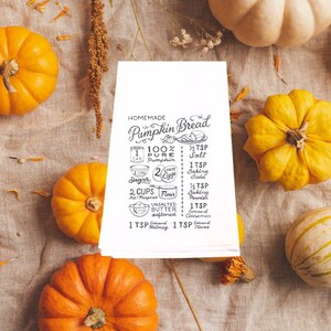 May include: A white recipe card with the words "Homemade Pumpkin Bread" and a list of ingredients, surrounded by various orange pumpkins and gourds on a tan fabric surface. The recipe includes ingredients like pumpkin, sugar, flour, and spices.