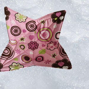 May include: A pink, star-shaped quilted hot pad with a floral and geometric pattern. The design includes brown, green, and pink flowers, hearts, and circles. The edges are trimmed in brown.