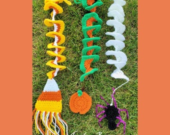 May include: Crocheted Halloween decorations including a candy corn broom, a pumpkin, a spider, and spiral garlands in green, white, and orange. The decorations are displayed on green grass. The text "Traverse Bay Crochet" is visible.