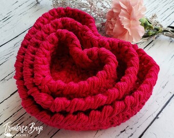 May include: A vibrant, hot pink crocheted heart-shaped basket. The basket is made of thick yarn and has a textured appearance. A pink carnation and silver decorative sprigs are in the background. The text "Traverse Bayx Crochet" is in the bottom left corner.
