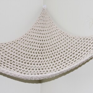 May include: Off-white, crocheted hammock-style storage net, suspended from a corner. The net features a diamond-shaped pattern and a thick, textured border. The hammock is designed to be mounted on a wall, offering a space-saving storage solution.