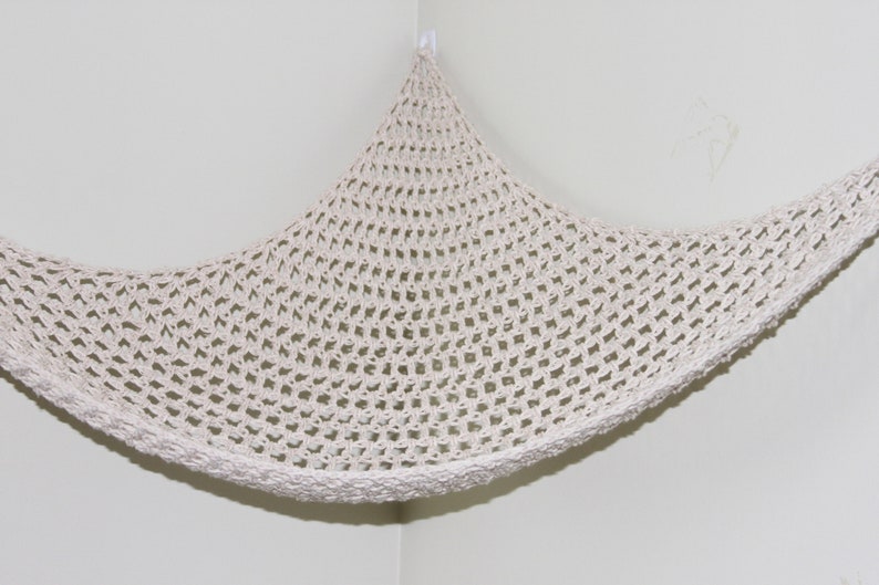 May include: Off-white, crocheted hammock-style storage net, suspended from a corner. The net features a diamond-shaped pattern and a thick, textured border. The hammock is designed to be mounted on a wall, offering a space-saving storage solution.