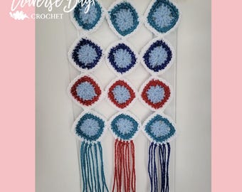 May include: A handmade crochet wall hanging featuring a grid of blue, red, and white diamond-shaped motifs. The wall hanging is suspended from a wooden dowel with hanging strings. The text "Traverse Bay Crochet" is in the upper left corner.