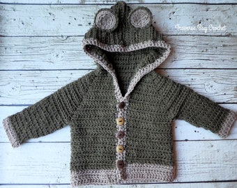 May include: A handmade, olive green crochet baby sweater with a frog hood. The hood features two circular ears. The sweater has a light brown trim and wooden buttons. The text "Traverse Bay Crochet" is visible in the upper right corner.