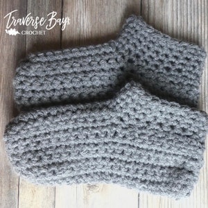 May include: A pair of gray crocheted slippers. The slippers are made of a thick yarn and have a textured appearance. The slippers are designed to cover the foot and ankle. The text "Traverse Bay Crochet" is in the upper left corner.