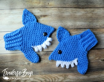 Puede incluir: Dos artículos de ganchillo azul con forma de tiburón, con dientes blancos y ojos de botón negros. Los artículos están sobre una superficie de madera. El texto "Traverse Bays Crochet" es visible en la esquina inferior izquierda.