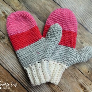 May include: A pair of handmade crochet mittens in shades of pink, red, gray, and white. The mittens feature a striped design with a pink top, red middle, and gray palm. The cuffs are white. The logo "Traverse Bay Crochet" is in the bottom left corner.