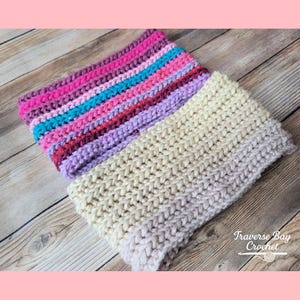 Peut inclure: Un col en crochet fait à la main en deux sections. Une section présente des rayures horizontales dans les tons de rose, de violet et de sarcelle. L'autre section est de couleur crème unie qui s'estompe en gris clair. Le texte "Traverse Bay Crochet" est dans le coin inférieur droit.