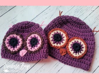 May include: Two purple crochet owl hats. The hats feature large, round eyes with brown, pink, and black details. The hats have small, pointed ears and a triangular beak. The hats are handmade and suitable for children.