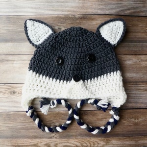 May include: A handmade crochet wolf hat. The hat is dark grey with a white face and ears. Black buttons form the eyes and nose. Braided ties with tassels hang from the ear flaps. A fun, cosy accessory.