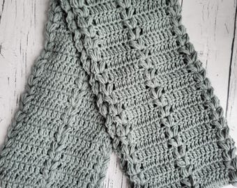 Può includere: Una sciarpa grigia all'uncinetto fatta a mano con un motivo a punto strutturato. La sciarpa è rettangolare, con un bordo dettagliato e un design ripetuto. Il testo "Traverse Bay Crochet" è visibile nell'angolo in basso a sinistra.