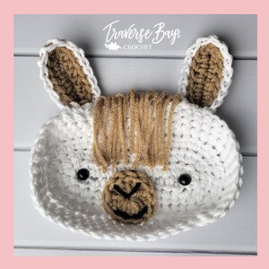 May include: A white crocheted llama-shaped basket with brown accents. The basket features a detailed face with black eyes and a brown nose. The ears and mane are also brown. The text "Traverse Bay Crochet" is visible.