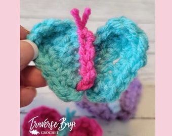 May include: Crocheted butterfly in shades of turquoise and pink. The butterfly has a pink center and antennae. The image also shows other crocheted butterflies in pink and purple. The text "Traverse Bay Crochet" is visible.