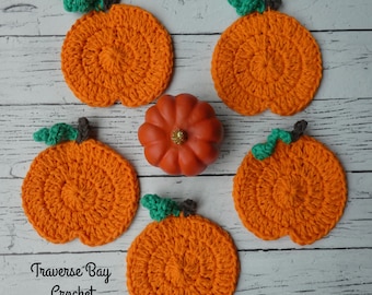 May include: Five orange crocheted pumpkin coasters with green and brown accents, arranged around a small orange pumpkin with a gold glitter top. The text "Traverse Bay Crochet" is visible on the bottom left. The coasters are on a white wooden surface.