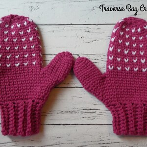 May include: A pair of magenta crochet mittens with white heart-shaped accents. The mittens are handmade, featuring a ribbed cuff and a textured stitch pattern. The text "Traverse Bay Crochet" is visible in the upper right corner.