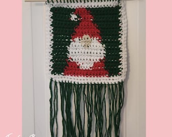 May include: A handcrafted crochet wall hanging with a Santa Claus motif. The design features a red hat and beard set against a green backdrop, bordered with white and green crochet. Green fringe hangs from the base. The text "Traverse Bays Crochet" is at the bottom.