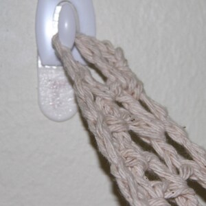 May include: A close-up of a white adhesive hook on a wall, holding a beige macrame plant hanger. The macrame is made of thick, woven cotton threads, creating a net-like structure. The hook is rectangular with a rounded button.