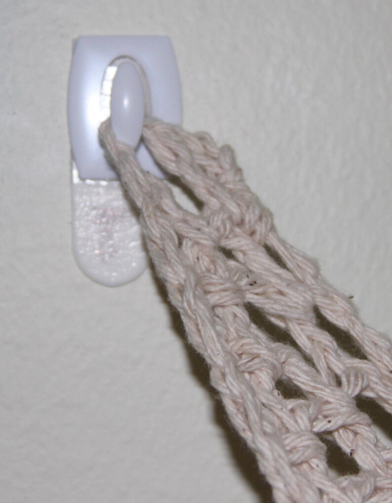 May include: A close-up of a white adhesive hook on a wall, holding a beige macrame plant hanger. The macrame is made of thick, woven cotton threads, creating a net-like structure. The hook is rectangular with a rounded button.