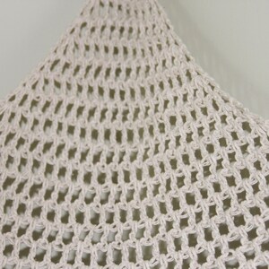 May include: Close-up of a cream-colored, open-weave macrame. The knotted pattern creates a net-like texture. The macrame is hanging against a neutral background, showcasing its intricate design and craftsmanship. This item is suitable for home decor.