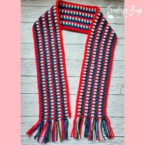 May include: A handmade crochet scarf in red, white, and blue. The scarf has a striped pattern with red borders and fringe. The text "Traverse Bay Crochet" is in the upper right corner.