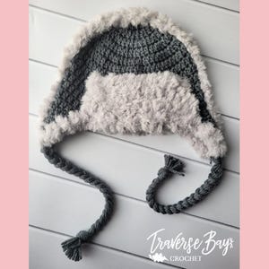 May include: A handmade crochet hat with a gray and light gray color scheme. The hat features a furry trim and ear flaps, with braided ties ending in tassels. The hat is designed for warmth and style.