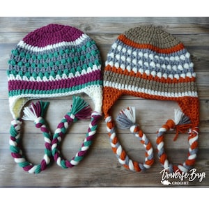 May include: Two handmade crochet hats with ear flaps and braided ties. One hat is mainly purple, teal, and white, the other is brown, orange, and white. The hats are displayed on a wooden surface.