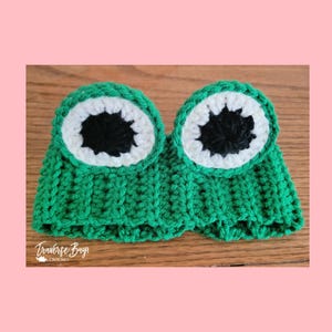 May include: A handmade crochet frog eye mask in bright green, white, and black. The mask features two large, round eyes with white and black details. The text "Traverse Bay Crochet" is visible in the lower left corner.