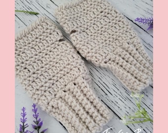 May include: A pair of beige crochet fingerless gloves. The gloves are made with a textured stitch and have a ribbed cuff. The gloves are displayed on a white wooden surface with flowers and the text "Traverse Bay Crochet".