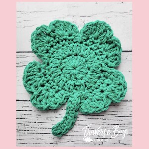 May include: A handmade, green crochet shamrock with four leaves. The clover is made of thick yarn and has a textured appearance. The stem is also crocheted and extends from the bottom of the clover. The background is a white wooden surface.