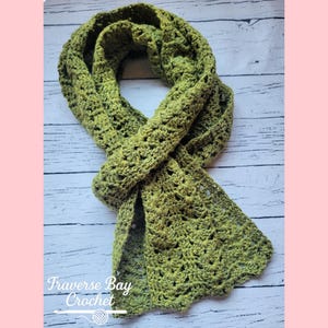 May include: A handmade, olive green crochet scarf. The scarf is made with a textured stitch pattern, and the ends have a scalloped edge. The text "Traverse Bay Crochet" is visible in the lower left corner.