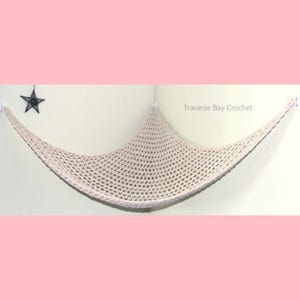 May include: A beige, crocheted cat hammock suspended from a corner. The hammock is made of a light-colored yarn with a net-like pattern. The text "Traverse Bay Crochet" is visible. A black star decoration hangs on the wall.