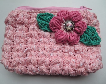 Puede incluir: Un monedero rosa tejido a mano con un patrón de puntada texturizada. Presenta una flor rosa con un centro decorativo y hojas verdes. El bolso tiene un cierre de cremallera rosa.
