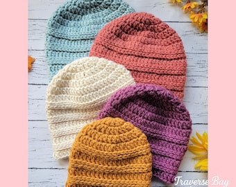 May include: A collection of six handmade crochet beanies in various colors, including blue, cream, peach, purple, and mustard yellow. The hats are arranged on a light wood surface. The text "Traverse Bay Crochet" is visible in the bottom right corner.