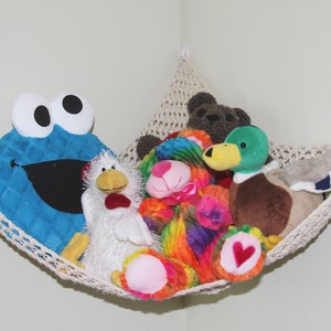 May include: A cream-colored macrame hammock filled with plush toys. A large blue Cookie Monster toy is on the left, with a white chicken and other colorful stuffed animals. A brown teddy bear and a green and brown duck are also visible.