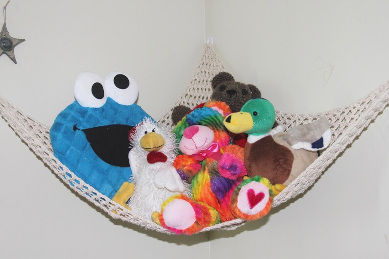 May include: A cream-colored macrame hammock filled with plush toys. A large blue Cookie Monster toy is on the left, with a white chicken and other colorful stuffed animals. A brown teddy bear and a green and brown duck are also visible.