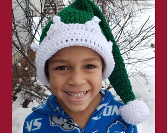 May include: A green and white crochet elf hat with a long, pointed tip and a white pom-pom. The hat features a white, scalloped brim. The hat is handmade and suitable for children. The text "Traverse Bay Crochet" is visible.
