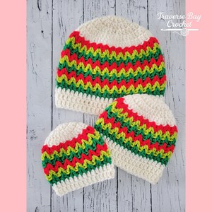 May include: Three crocheted beanies in varying sizes. The hats feature a cream-colored base with a zig-zag pattern in red, green, and yellow. The hats are arranged on a light wood surface.